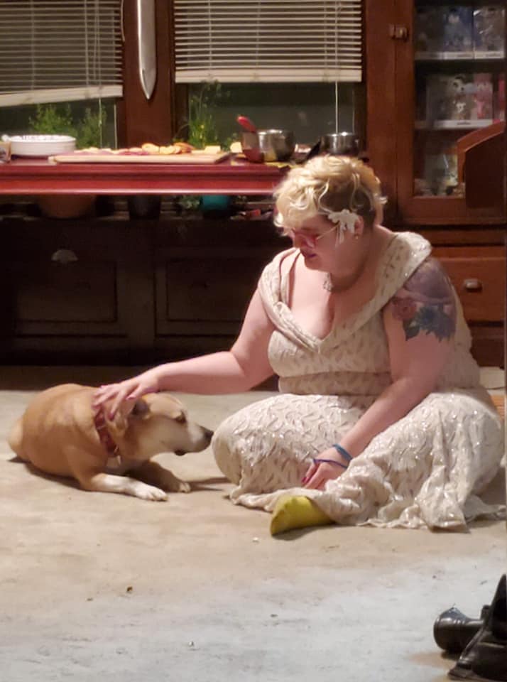 Caitlin is dressed for a 20s themed New Years Eve party in a white glittery gown. They are petting a dog, because that is what Caitlin does at a party.