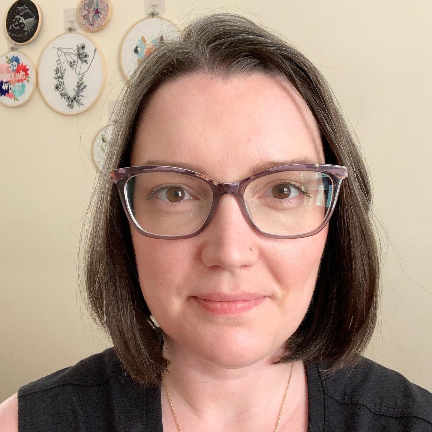 Anne-Marie is smiling softly at the camera, facing directly forward. She has chin length brown hair and is wearing purple glasses. Behind her are several embroidery pieces she has made, hanging on her wall.