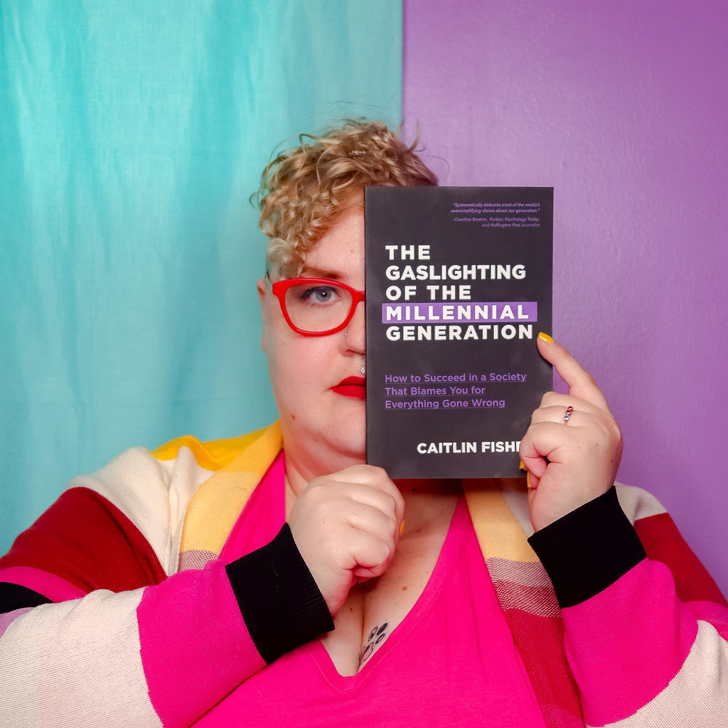 Caitlin holding a copy of their book, The Gaslighting of the Millennial Generation, in front of their face. It looks very edgy and cool.