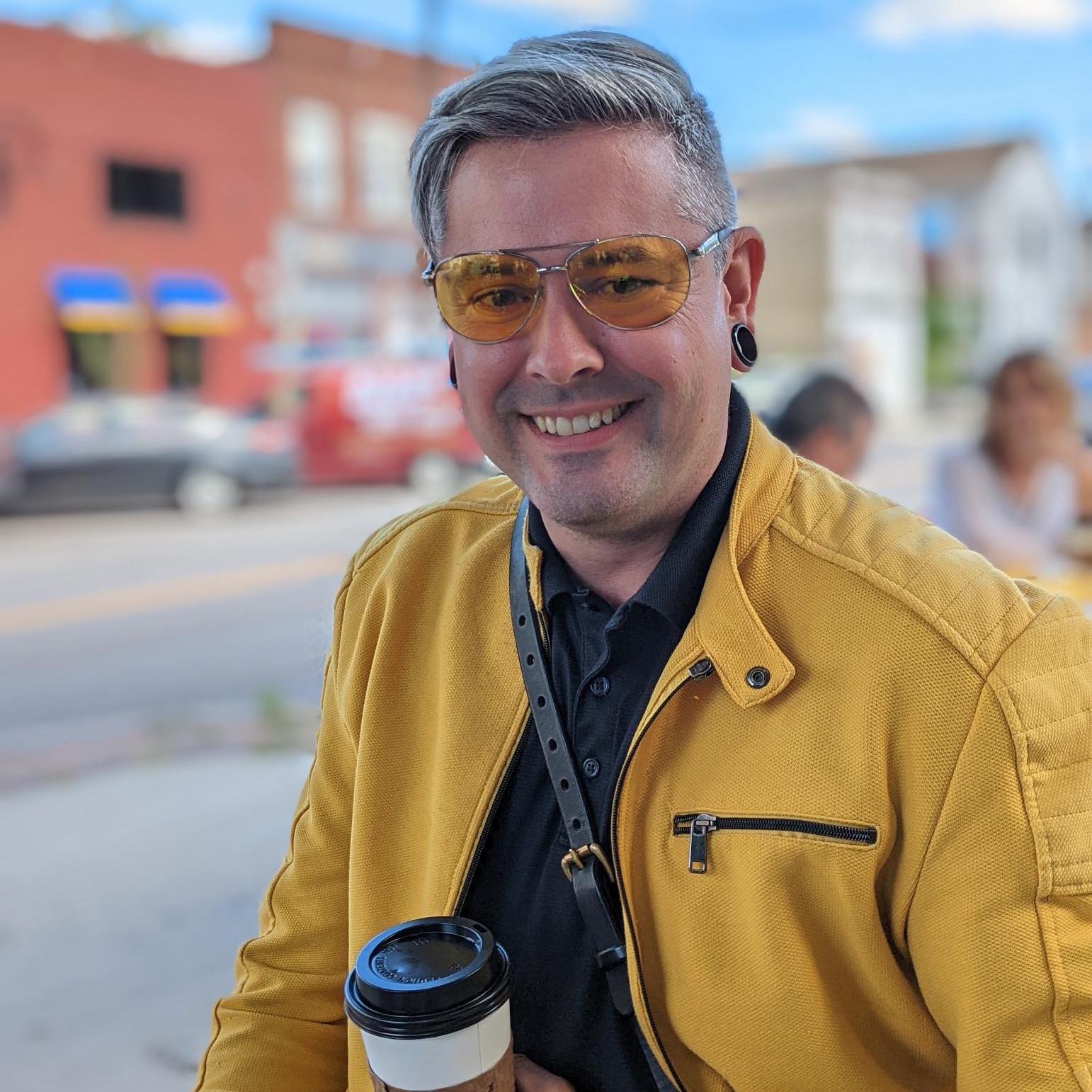 Tristan is looking at the camera with a smile on their face, wearing yellow tinted sunglasses, a yellow motorcycle style jacket, and a black button-up shirt. They have a leather strap for a bag across their body, gauged ear piercings, and they're holding a cup of coffee.