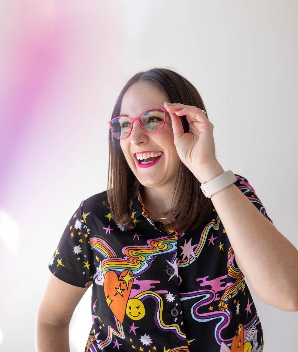 Kristy is smiling brightly off-camera and holding their hot pink glasses up in front of their eyes. They're wearing a truly queer button-up shirt with rainbows, hearts, stars, and lots of pink blobs, like a galaxy of joy in shirt form.