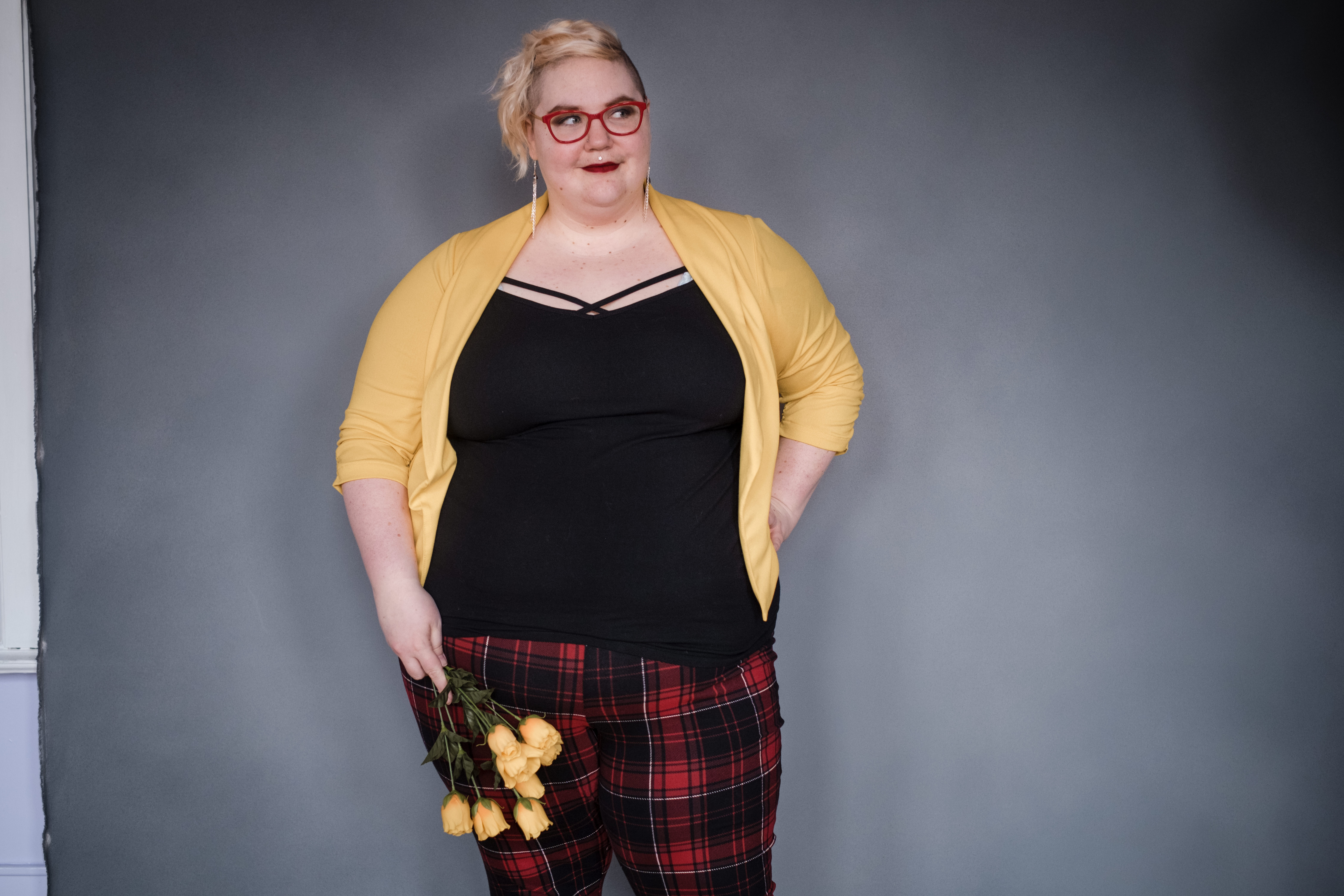 Caitlin is smiling and looking to the side, wearing a black tank top, yellow blazer, and red plaid pants. They have yellow tulips in one hand, and the other hand is on their hip. 