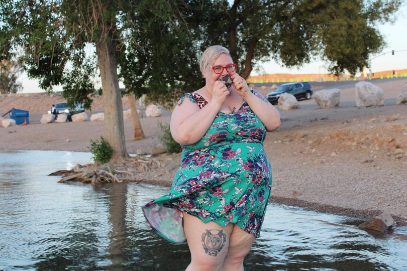 Caitlin wading in the Colorado River close to the bank. They are holding up a rock to their face with a happy expression you can see in their eyes. They're wearing an above-the-knee sundress that's blowing in the wind and you can see a tattoo of their cat on their right thigh.