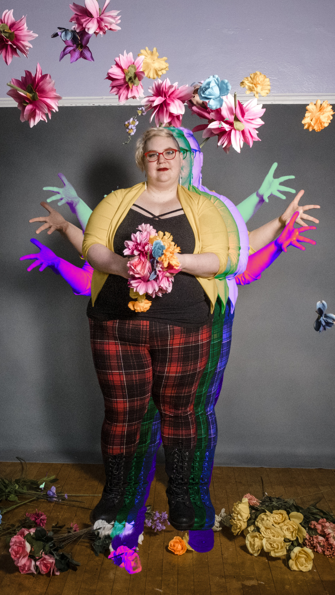 An edited photo of Caitlin throwing fake flowers into the air. The image of them throwing the flowers has been duplicated and there is a green filtered cutout and purple filtered cutout of their hands coming from behind their body so it looks like they have lots of arms. In front of the throwing arms posture is another image of Caitlin superimposed, with a handful of flowers before throwing them. There are also two duplicates of Caitlin in this standing/waiting posture edited behind their body. Overall it looks like six versions of Caitlin caught between two moments in time, when they held the flowers and when they threw the flowers. 