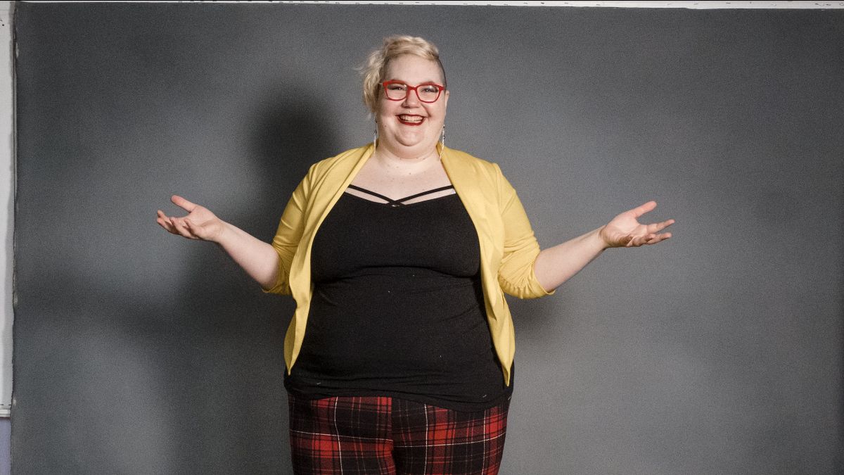 Caitlin is smiling directly at the camera with their arms spread in a questioning gesture